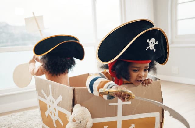 Zwei Kinder in der Kita sitzen in einem Piratenschiff aus einem Pappkarton, tragen Piratenhüte und spielen Pirat.