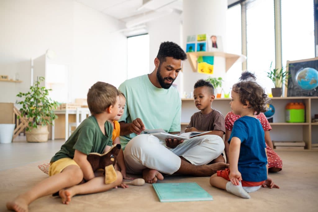 Ein Tagesvater sitzt im Kreis mit einer Gruppe von Kindern auf dem Boden und liest ihnen aus einem Buch vor.