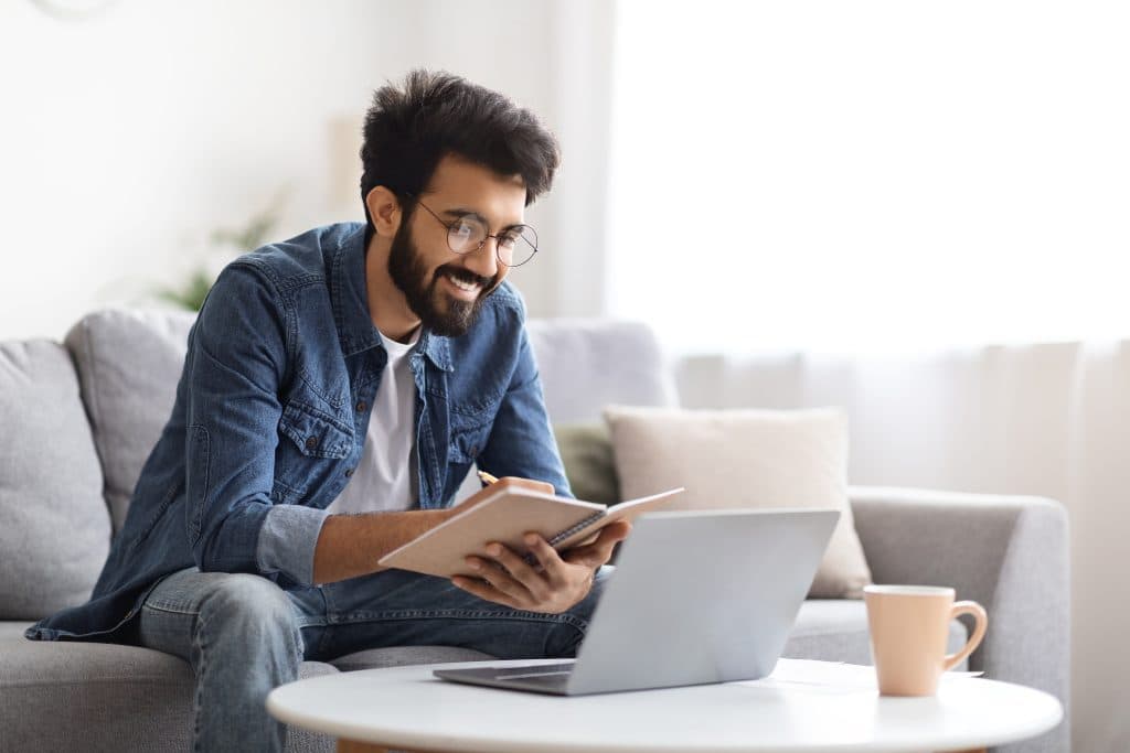 Erzieher mit Brille und Bart sitzt auf dem Sofa mit Laptop und Notizbuch. Er lernt für seinen Fernkurs.