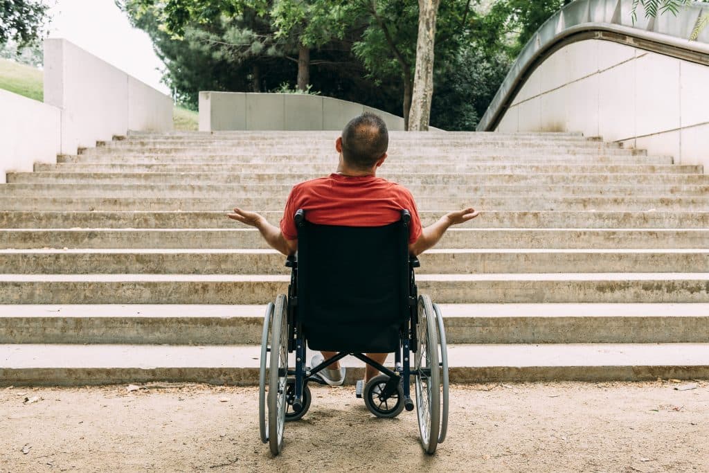 Ein Mann im Rollstuhl sitzt vor einer langen Treppe im Freien und hebt fragend die Arme – keine barrierefreie Zugangsmöglichkeit in Sicht.