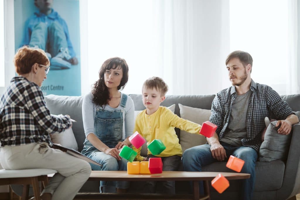 Eine Familie sitzt auf einer Couch und unterhält sich mit einem Therapeuten. Ein Kind spielt mit bunten Blöcken auf einem Tisch. Heller Raum mit einem Poster im Hintergrund.