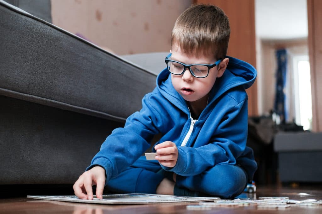 Ein Junge mit blauem Kapuzenpulli und Brille sitzt auf dem Boden und setzt konzentriert ein Puzzleteil zusammen.