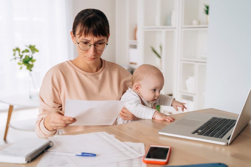 Frau mit Brille, die ein Dokument in der Hand hält, sitzt an einem Tisch mit einem Baby und greift nach einem Laptop. Auf dem Tisch liegen Papiere, ein Telefon und ein Notizbuch.