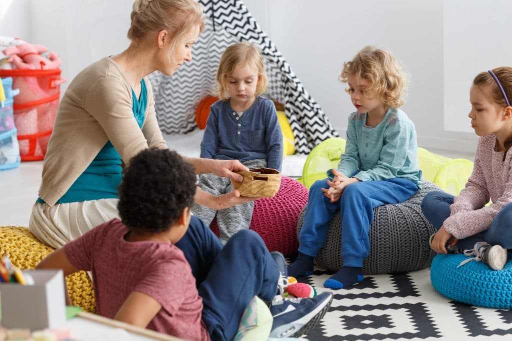 Pädagogische Fachkraft sitzt mit einer Gruppe Kinder auf Sitzkissen im Kreis.