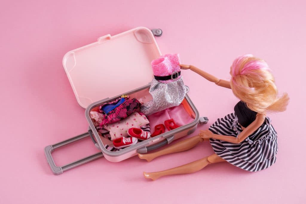 Puppe sitzt auf dem Boden mit geöffnetem Spielzeugkoffer und vielen Klamotten.