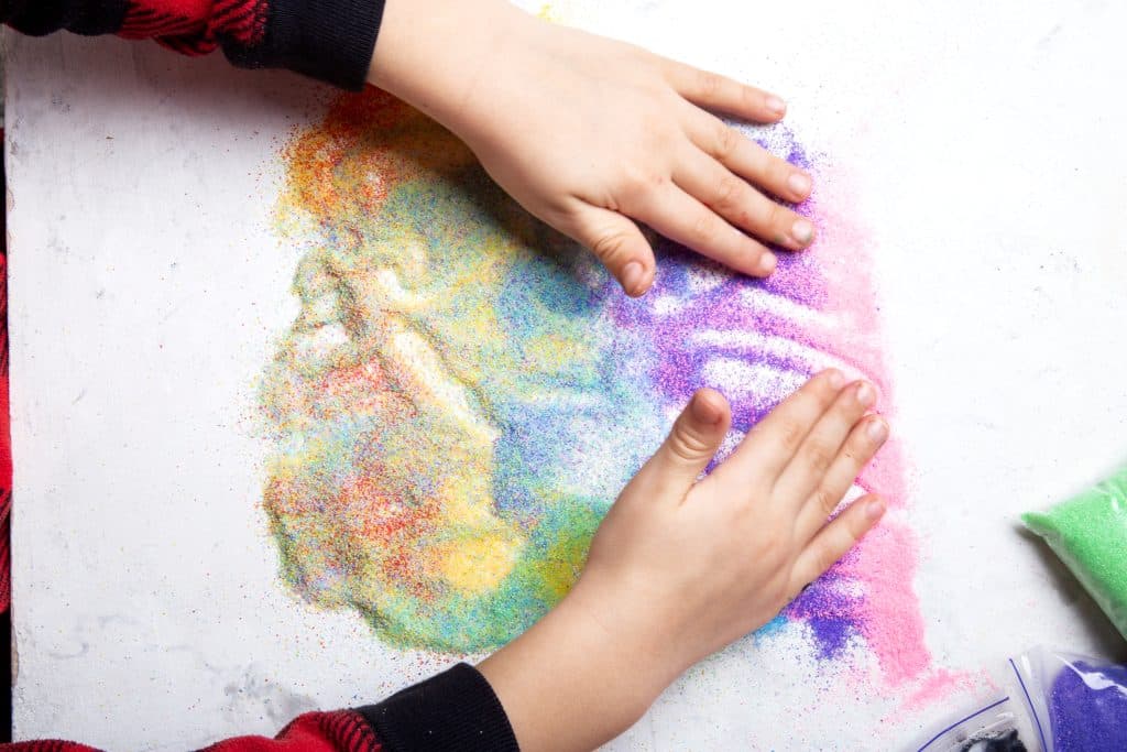 Kinderhände spielen mit buntem Sand auf einer weißen Platte.
