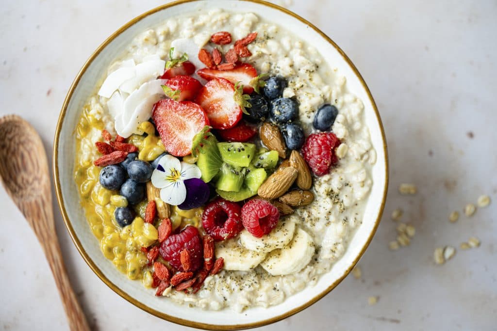 Porridge garniert mit Erdbeeren, Kiwi, Blaubeeren, Himbeeren, Mandeln, Bananenscheiben, Kokosflocken und essbaren Blüten.
