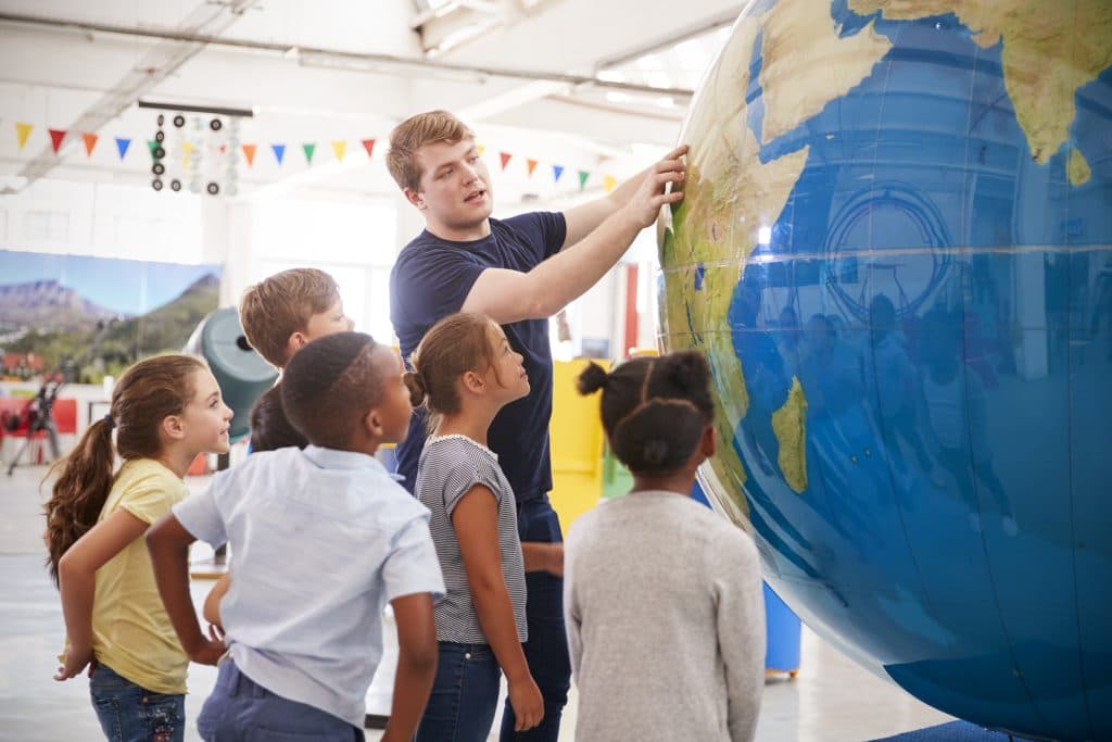 Erwachsener zeigt einer Gruppe Kinder einen Ort auf einem großen Globus in einer Ausstellung.