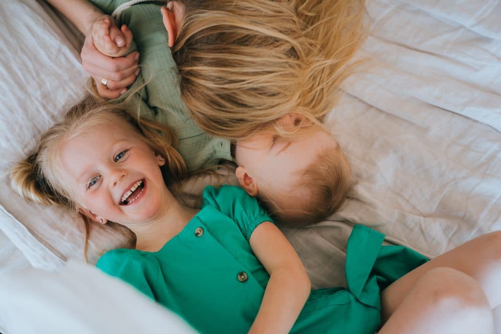 Zwei kleine blonde Kinder liegen in grüner Kleidung unter einer leichten Decke auf einem Bett – eines lacht, das andere kuschelt sich an seine Mutter.