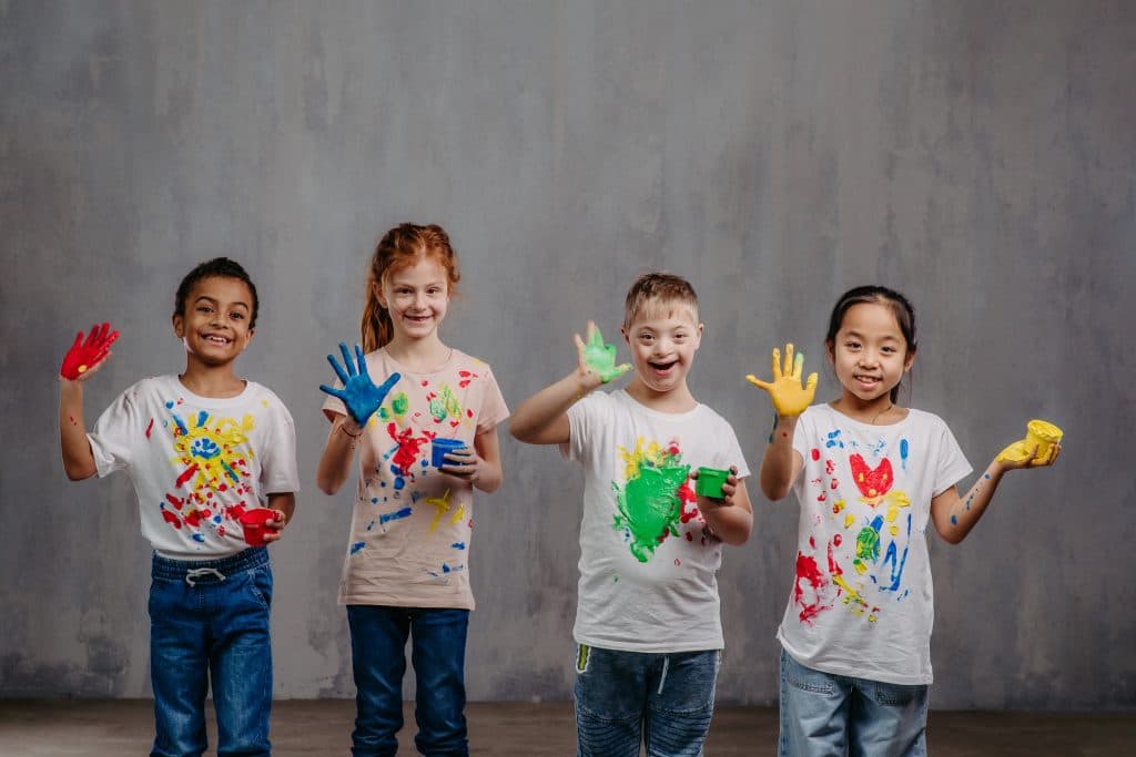Vier fröhliche Kinder mit bemalten Händen und T-Shirts stehen nebeneinander und zeigen ihre bunten Farben.