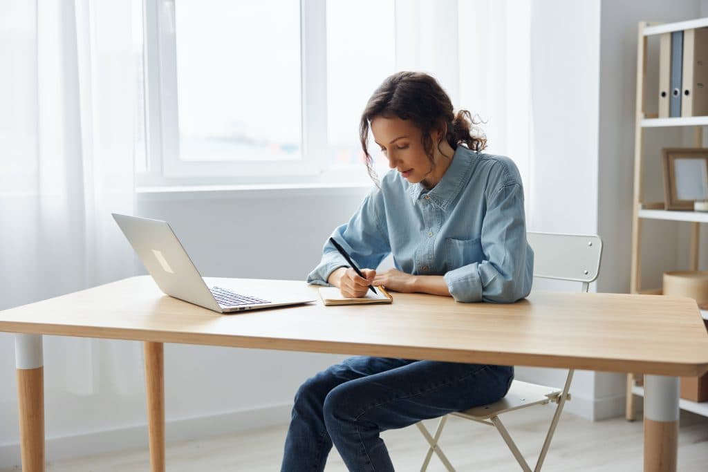 Eine Frau in einem Jeanshemd sitzt an einem Holztisch und schreibt in ein Notizbuch. Neben ihr ist ein Laptop in einem hellen, minimalistischen Raum aufgeklappt.
