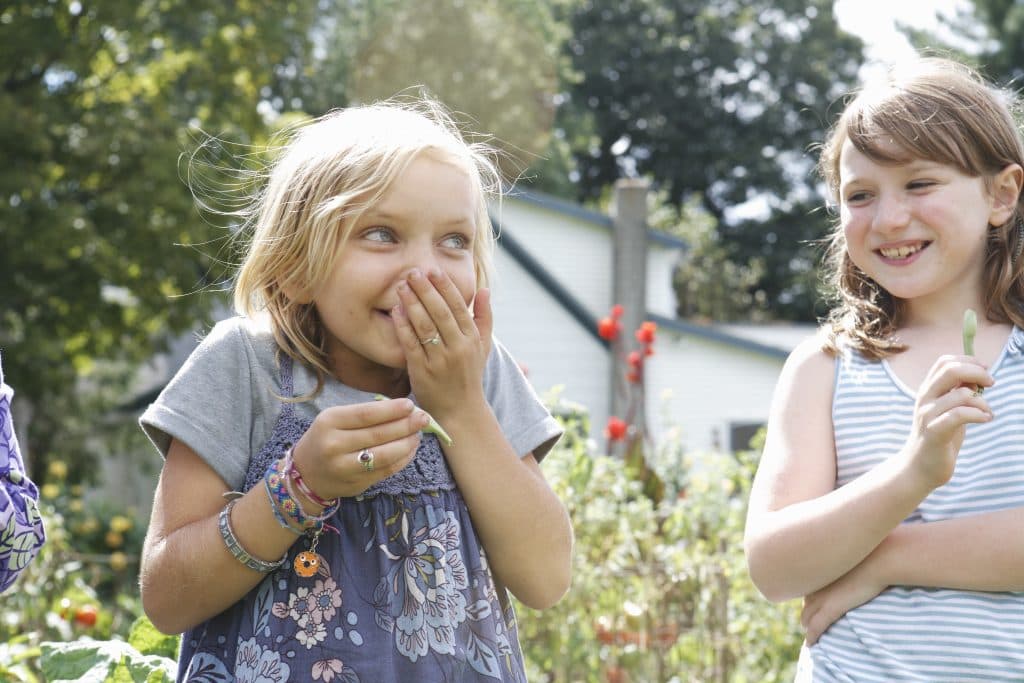 Zwei junge Mädchen lachen in einem Garten, eines hält sich spielerisch die Hand vor den Mund. Sie sind von Grünpflanzen und Blumen umgeben.