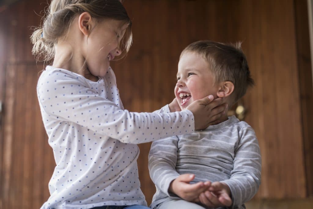 Zwei Kinder lachen fröhlich, eines hält sanft das Gesicht des anderen. Sie tragen legere Langarmshirts in einem warm beleuchteten Raum.
