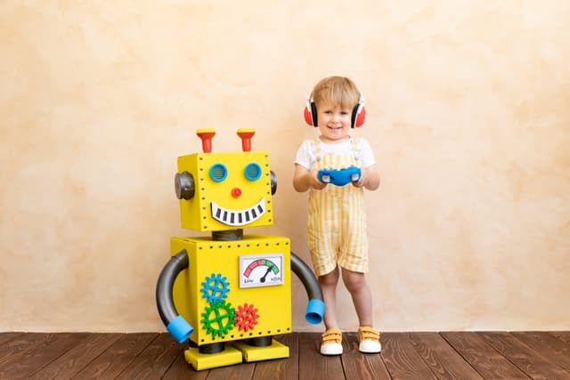 Kleines Kind mit Kopfhörern und Fernbedienung steht neben einem selbstgebastelten, fröhlich gelben Roboter.