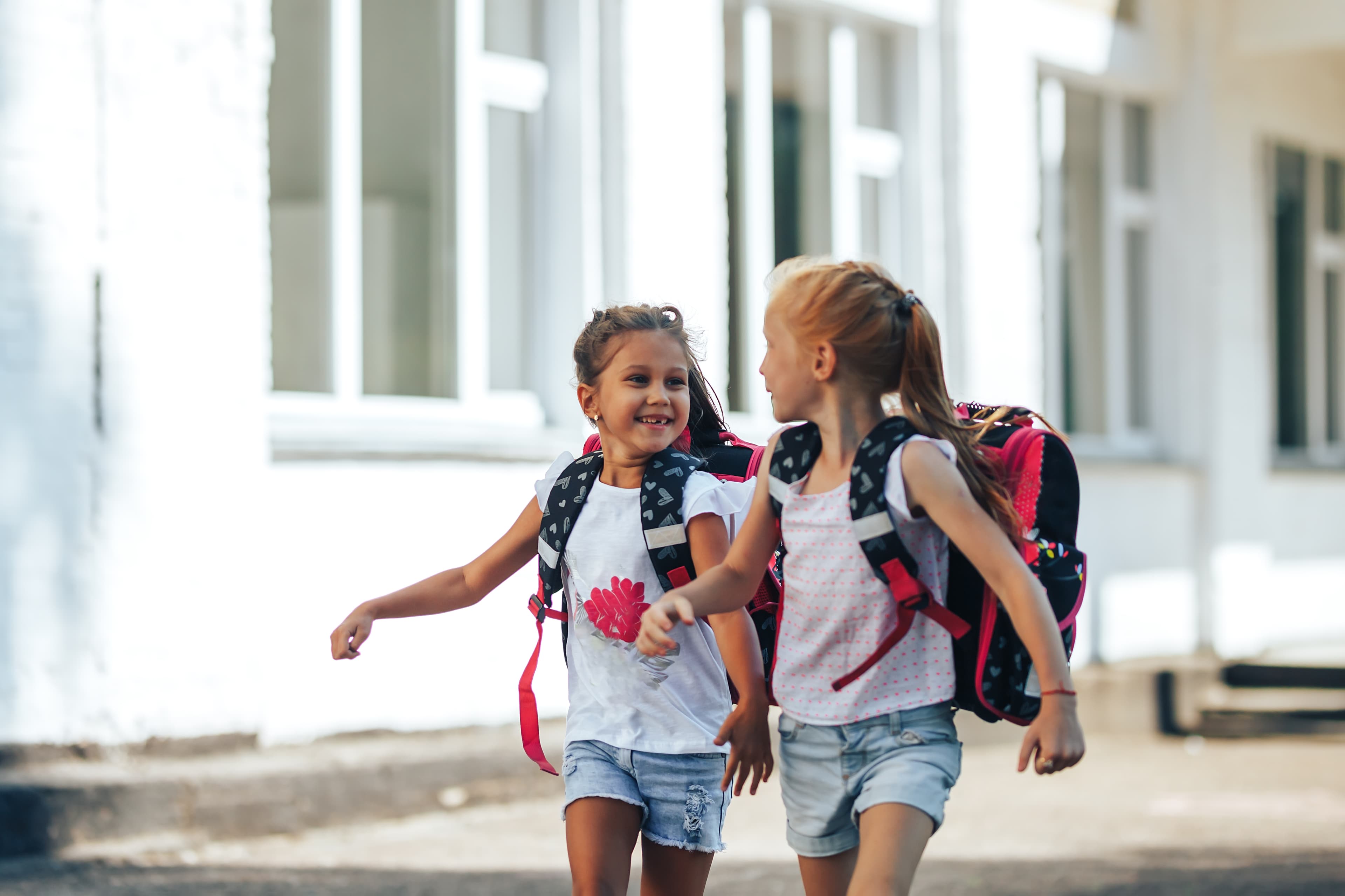 Zwei Grundschulkinder mit Schulranzen laufen lachend nebeneinander über den Schulhof.