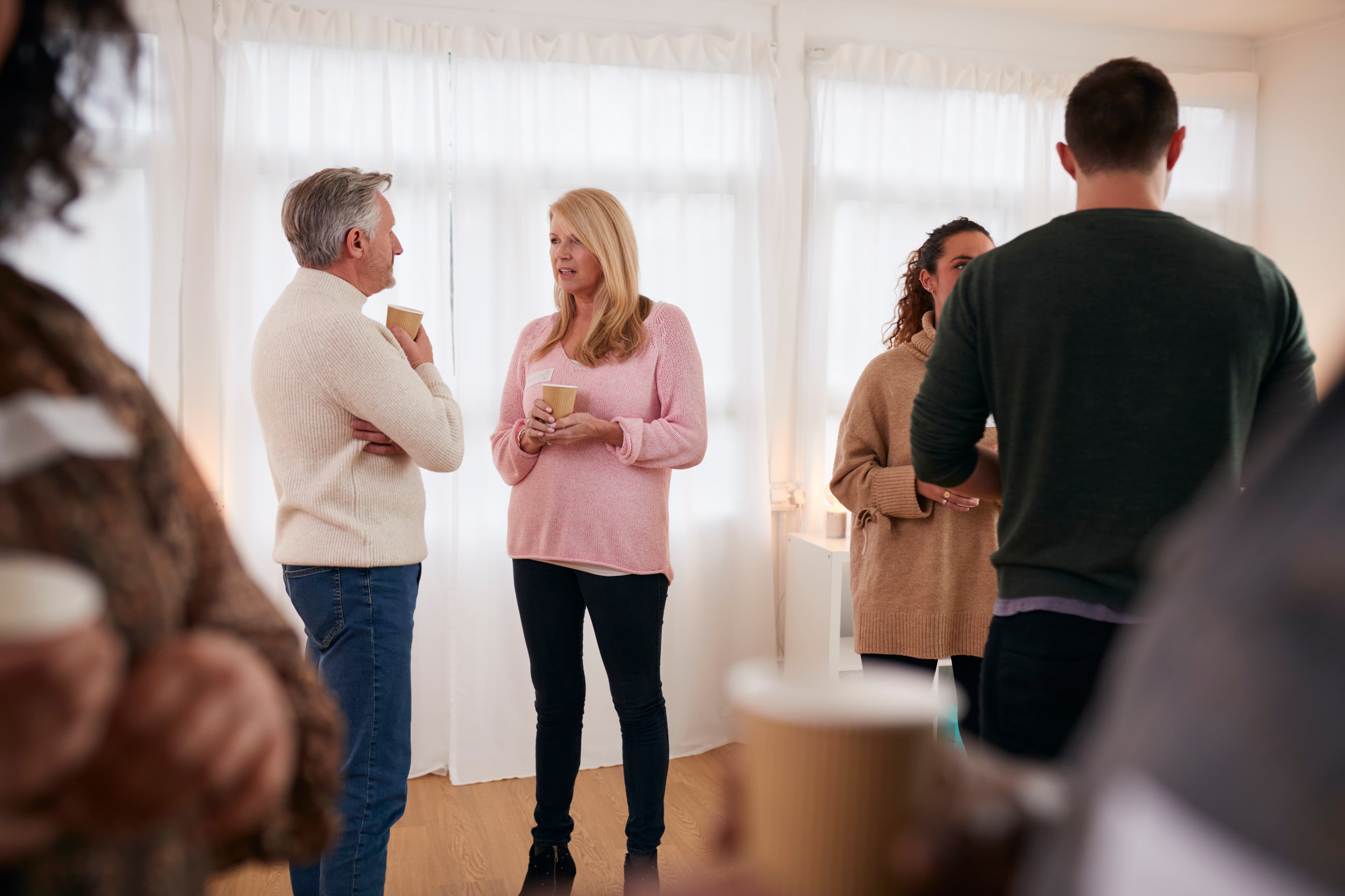 In einem hellen Raum unterhalten sich Menschen in Freizeitkleidung und halten Kaffeetassen in der Hand. Im Hintergrund sind weiße Vorhänge zu sehen.