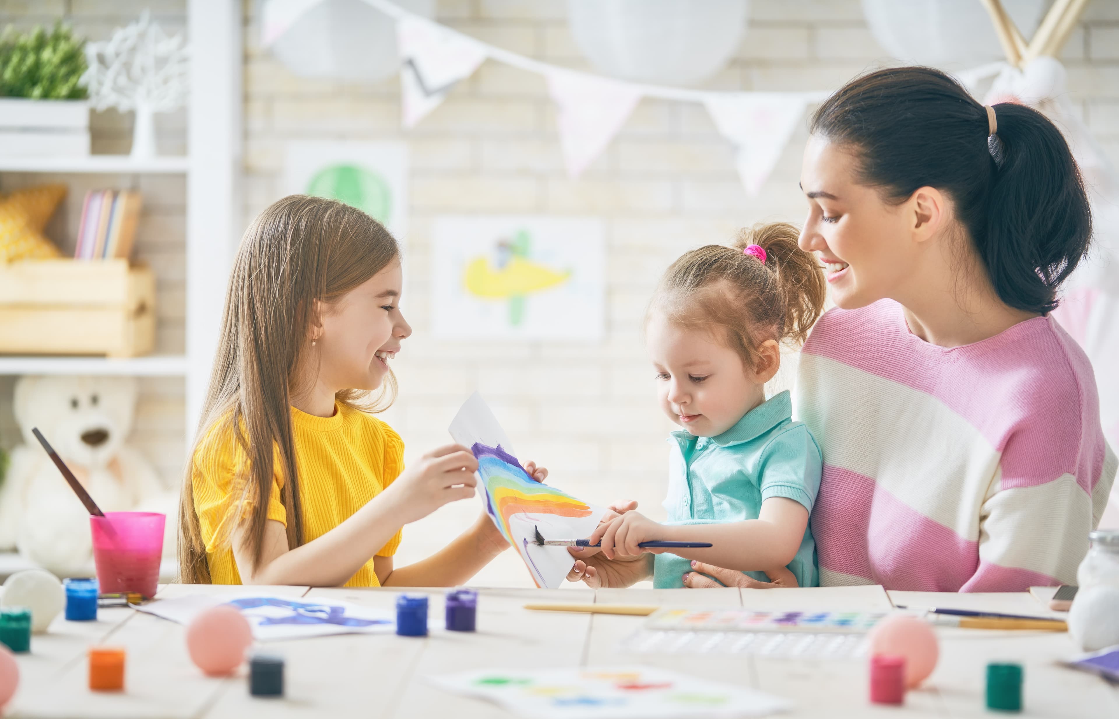 Zwei Kinder und eine Frau malen in einem hellen und freundlichen Raum an einem Tisch, umgeben von Malutensilien, einen bunten Regenbogen.