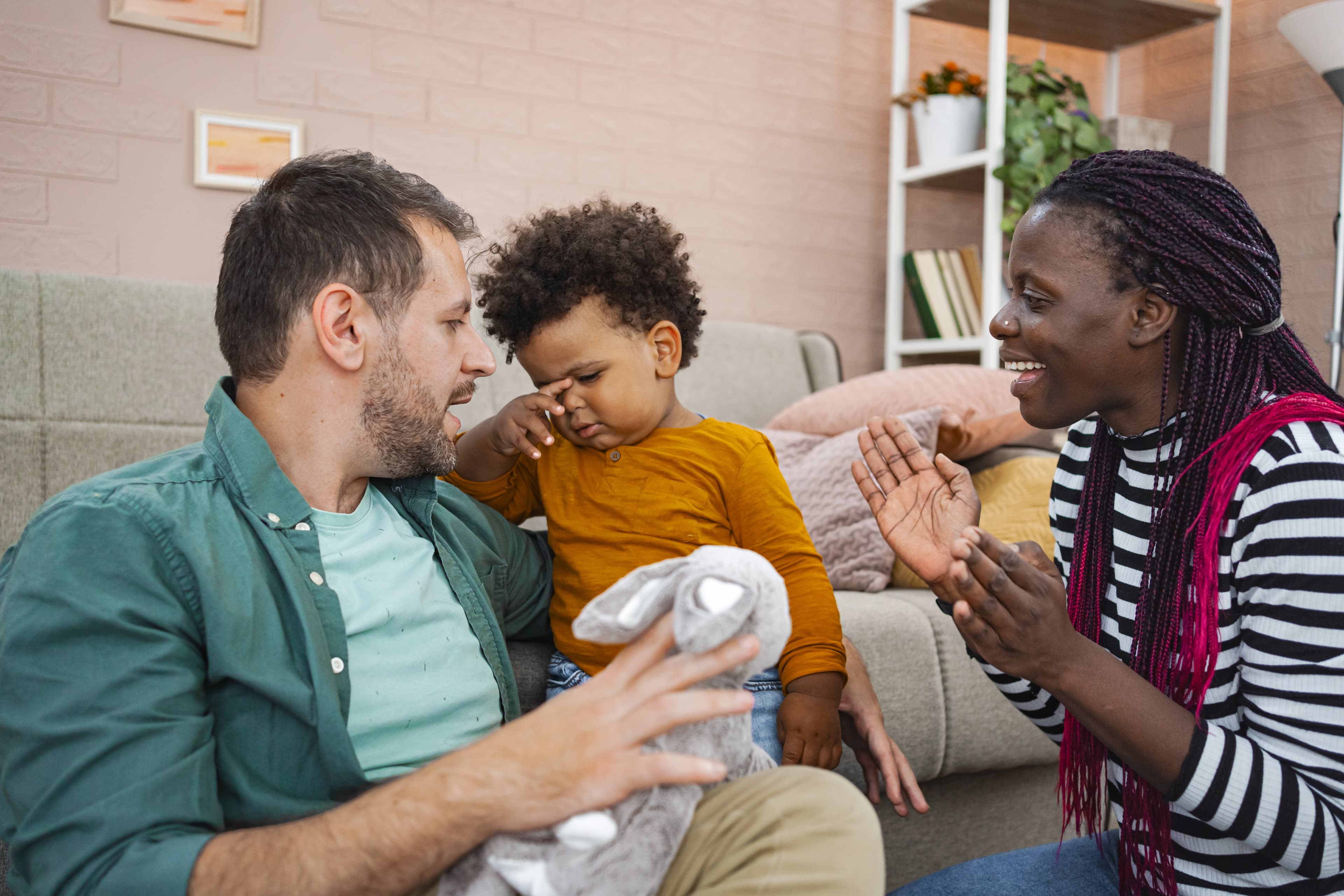 Ein Kind reibt sich die Augen, während es mit einem Mann und einer Frau zusammensitzt, die lächeln und mit dem Kind in einem gemütlichen Wohnzimmer interagieren.
