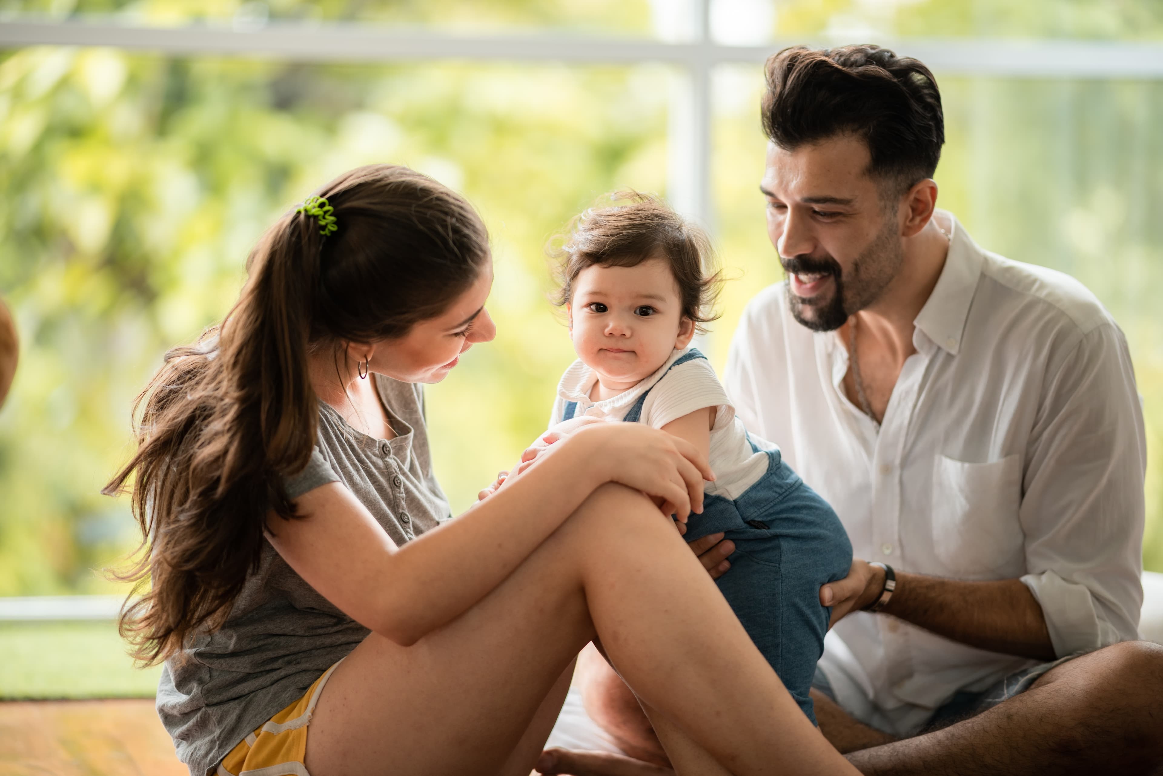 Eine dreiköpfige Familie sitzt zusammen in einem Raum, lächelt und beschäftigt sich mit ihrem Baby. Im Hintergrund ist ein Fenster zu sehen.
