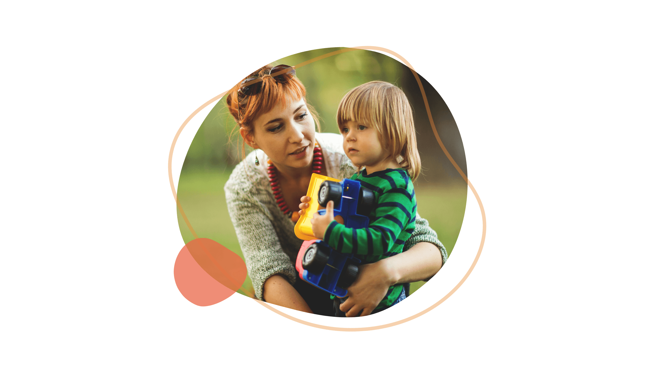 Frau mit roten Haaren hält ein Kind in einem Park, beide schauen auf einen Spielzeug-Lkw.