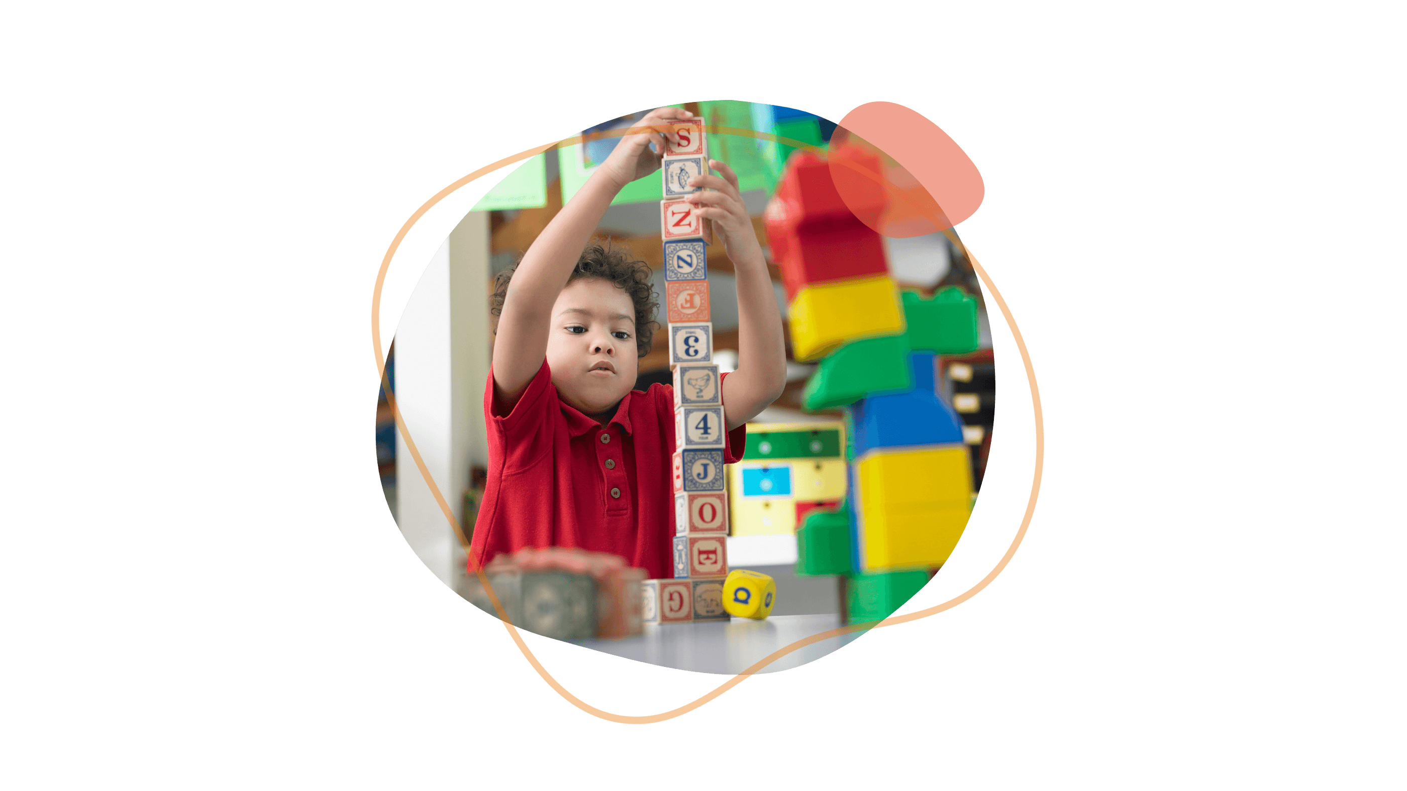 Ein Kind stapelt konzentriert Buchstabenbausteine in einem Klassenzimmer und streckt die Arme nach oben. Im Hintergrund sind bunte Spielsachen zu sehen.