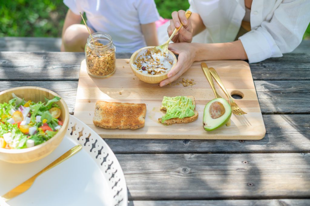 Erwachsene Person und Kind sitzen an einem Tisch. Vor ihnen liegt ein Brett und ein Tablett mit Broten, einer Avocado, Müsli und einer Schüssel Salat. Es geht um gesunde Ernährung von Kindern 