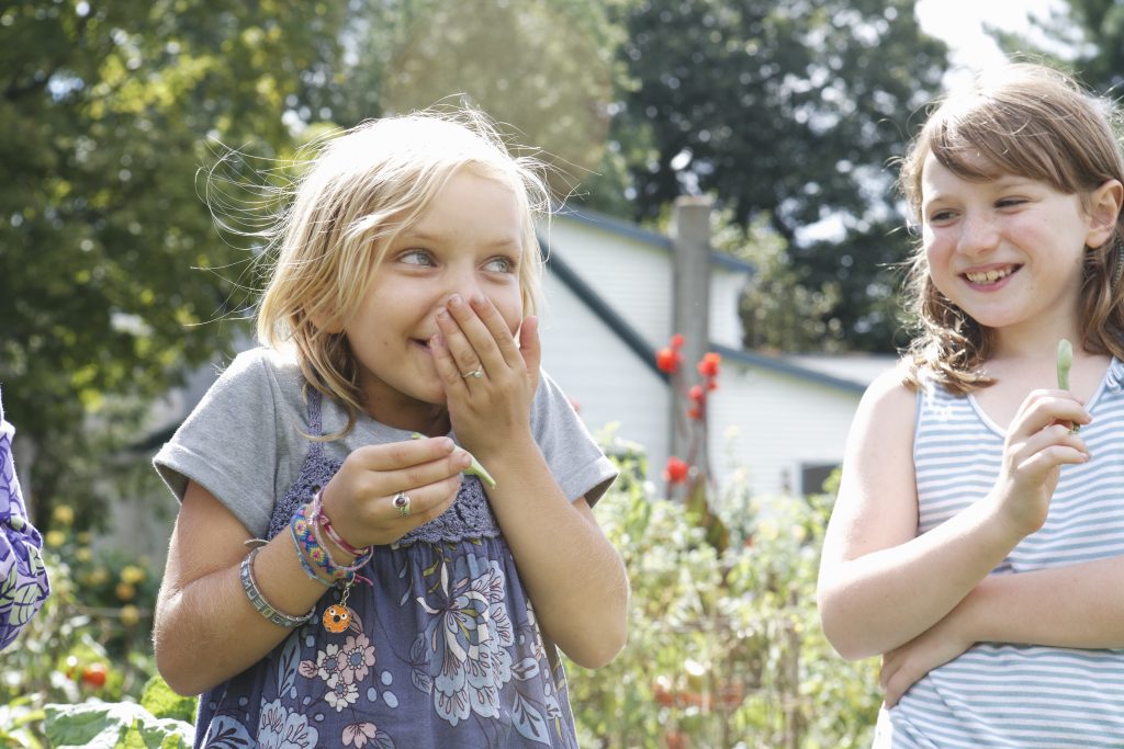 Zwei Mädchen stehen im Garten und lachen, das eine Mädchen hält sich dabei verschmitzt die Hand vor den Mund. Sexualpädagogik in der Kita ist immens wichtig!